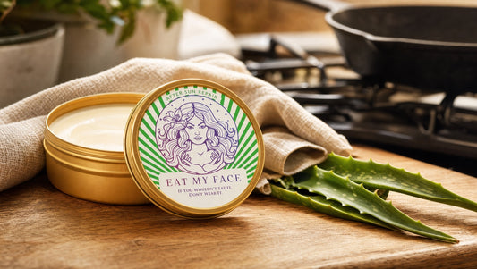 Open gold Eat My Face After Sun Balm tin with visible balm on a warm wooden kitchen counter beside a cast-iron skillet and fresh aloe leaf