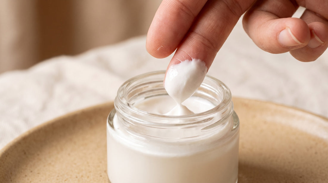Fingertips lifting a dollop of thick white mineral sunscreen from a glass jar