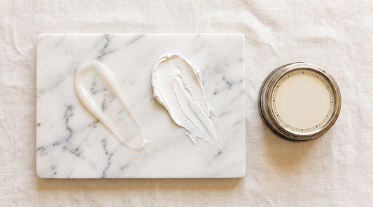 Two cream swatches on marble beside a kitchen timer, illustrating sunscreen or lotion first