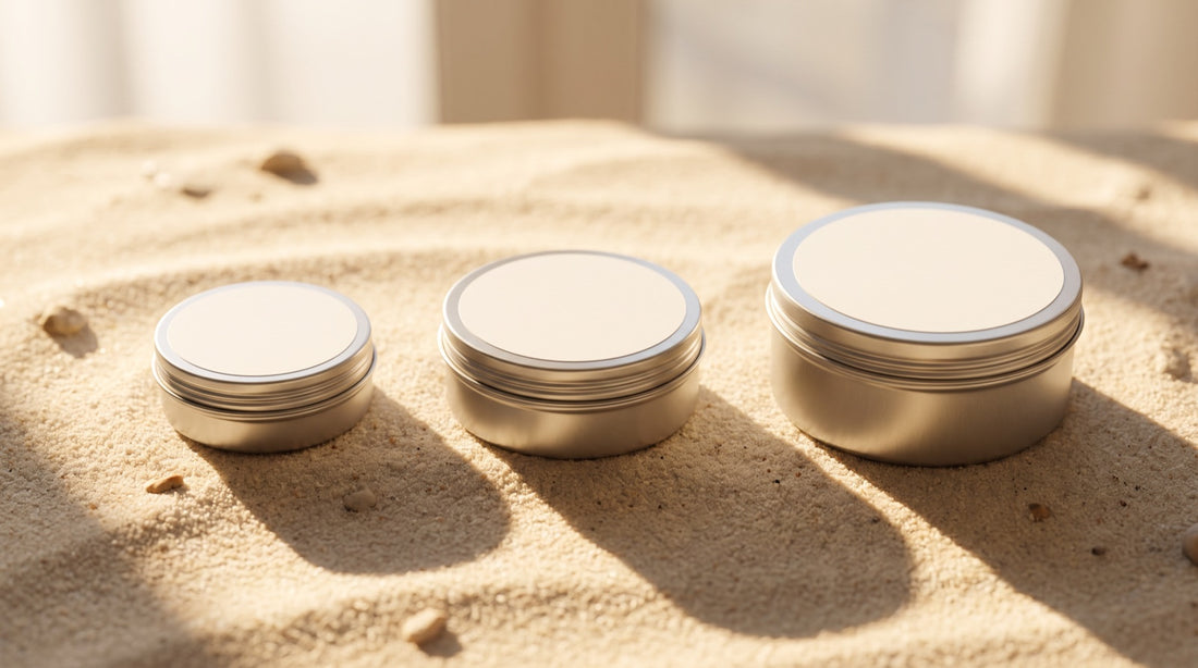 Three round metal tins of mineral sunscreen in increasing sizes lined up on bright sunlit sand