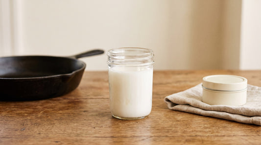 A jar of white beef tallow between a cast-iron skillet and a folded linen towel with a small cream tin
